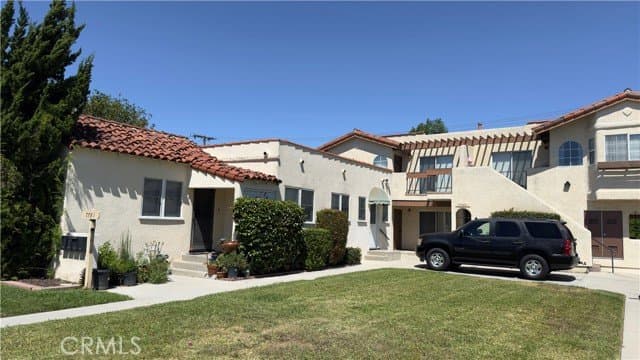 An exterior view of a multi-unit residential property with a front house featuring a red tiled roof, a lawn, and a two-story rear building with an external staircase, balconies, and a dark SUV parked in front. The address '7781' is visible on a sign by the front house.