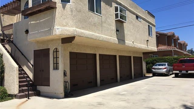 A beige multi-story triplex building with three brown garage doors, an external staircase leading to upper units, and a concrete driveway with a silver car and a red truck parked.