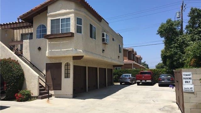 A beige multi-family building with a red tile roof and four brown garage doors on the ground level, beside a wide concrete driveway with parked cars.