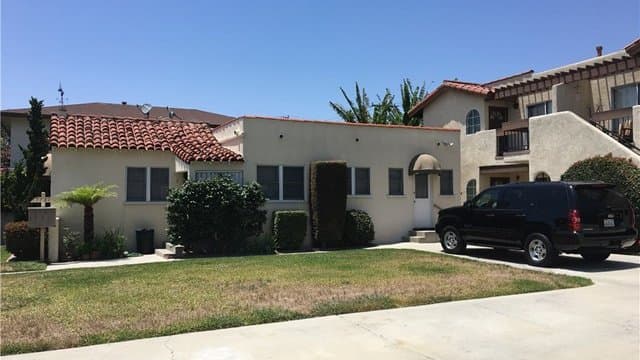 Front view of a cream-colored house with a red tile roof and a grassy yard, with an adjacent multi-unit building and a black SUV in the driveway under a clear blue sky.